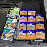 Photo showing various packs and spindles of blank DVDs, CDs, and one audio cassette tape arranged on black surface.