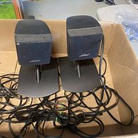 Front view of the two Bose small cube speakers on black stands showing cables and fabric grills with Bose logo.