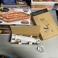 Photo showing Gotham Bacon Bonanza copper bacon rack box, Pampered Chef cookie press box, and Procter Silex electric knife in box on a table