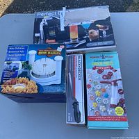 Photo showing box of Perfect Drink Pro smart drink scale and dispenser, box of Great American Steakhouse onion machine, Black & Decker knife sharpener box, and Nordic Ware cookie press box.