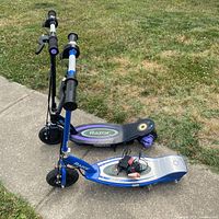 Two Razor electric scooters, one blue and silver, one black and purple, positioned side by side outdoors on sidewalk and grass.