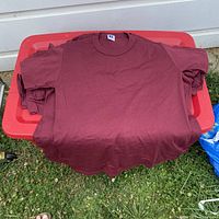 Lot of 21 maroon red short sleeve vintage t-shirts displayed folded over a red bin outdoors on grass.
