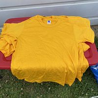 Nine yellow t-shirts stacked together showing the front side, plain with no prints or graphics, on a red surface outdoors.