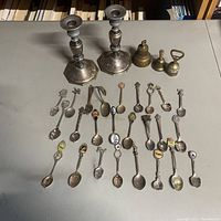 A set of 22 mini collectible souvenir spoons laid on a table with three small bells and two metal candlesticks, clear view of design and condition.