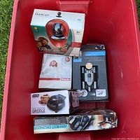 Lot viewed inside red plastic tote; Logitech webcam box visible, Miele vacuum bags, binoculars box, mini griddle maker box, arm rest organizer box