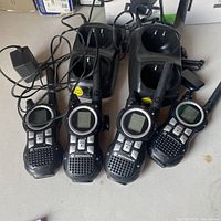 Four Motorola walkie talkies lying side by side with two charging docks and power adapters, showing front view with buttons, screens, and antennas.