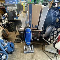 Front view of Oreck Magnesium vacuum cleaner upright with blue fabric bag and silver base, surrounded by household items.
