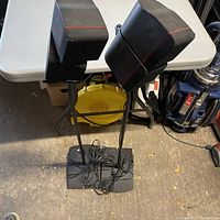 Pair of Bose cubed speakers on black metal stands with cables coiled at the base, sitting on a concrete floor next to a white table.