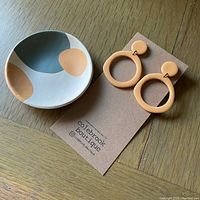 Photo showing polymer clay earrings on branded card alongside polymer clay trinket dish with abstract color pattern on wooden surface.