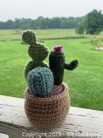 Crochet potted cactus plant with three green cactus shapes and a pink flower in a brown pot sitting on a railing outdoors with a green field background.