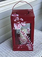 Front and side view of red metal candle lantern with hand painted pink and white flowers and leaves, showing the included white pillar candle inside.