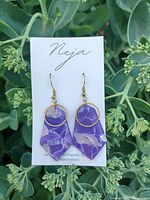 Photo of two handcrafted dangle earrings by Neja Creations, purple resin with white marbling and gold circular accents against green foliage background.