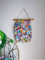 Front view of colorfully shaggy textile wall hanging suspended from a wooden dowel with string, showing vibrant mixed yarns and dimensions.