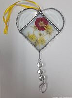 Front view of a hand-made stained glass heart with natural pressed flowers in the center and a yellow ribbon for hanging, along with a decorative glass bead dangle.