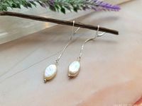 Photo of sterling silver threader earrings with oval freshwater pearls laid side by side on a pale surface with some lavender sprigs in the background.