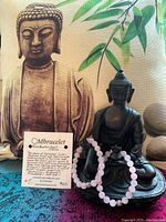 Two OMbracelets displayed on a Buddha statue with a handwritten description card about the Rose Quartz Heart Chakra bracelet.