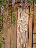 Macrame wall hanging displayed outdoors on wooden fence, surrounded by purple flowers, showing full vertical length and design