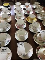 Top down view showing 6 assorted teacup and saucer sets with floral designs and gold trim, arranged on a wooden table.