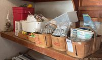 Photo shows wooden baskets filled with various hardware items such as screws, nails, switch plates and electrical boxes, located on a wooden shelf with a red plastic bin behind.