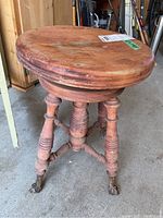 Vintage wooden piano stool showing turned spindle legs, round seat, and glass ball feet with casters on a concrete floor.