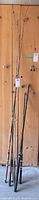 Full view of all six fishing rods leaning against wooden panel, showing condition and variety in handles and rod lengths.