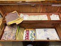 Collection of folded linens including polka dot apron, assorted fabrics, and packaged linens in wooden compartment