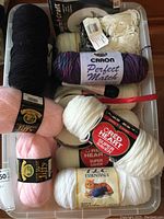 Plastic tote with various skeins of yarn including Caron Perfect Match, Red Heart Super Saver, TLC Essentials cat label, and Lion Brand Jiffy pink skeins.