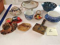 Overview of lot showing various ceramic teacups, bowls, carved figures, brass silent butler and other items on flat surface