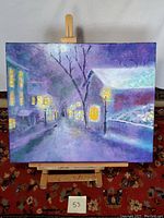 Frontal view of the oil painting on wooden easel showing the street light night scene with glowing windows and bare trees in a purple-toned impressionistic style.