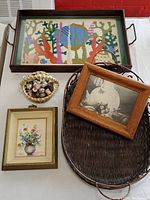 Top-down view of colorful rectangular needlepoint serving tray with metal handles, small basket of vintage sewing notions, miniature floral painting, framed pig cartoon photo, and oval wicker basket.