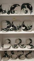 Sango Quadrille Black Lillies china displayed on shelves showing multiple dinner plates, bowls, teacups, saucers, cream and sugar bowl items with consistent floral design.