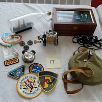 Top view of the lot on a white surface showing ship in a bottle, fishing reel, uniform patches, buttons, camping canteen with strap, and other small items.