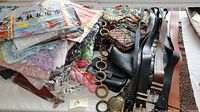 Overview of silk scarves in colorful and pastel shades stacked with some lace scarves sealed in plastic and visible purses and belts beneath