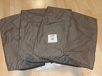 Three beige square Sunbrella outdoor pillowcases laid flat on a wooden floor, showing front surfaces and tags.