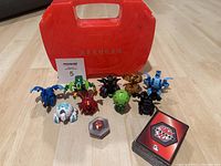 Nine Bakugan figures, game cards, hexagonal token, and red Bakugan case displayed together on floor.