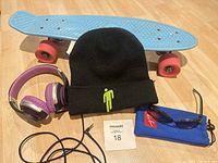 Full lot including blue Penny skateboard with pink wheels, black Billie Eilish beanie, purple and silver foldable headphones, and Ray-Ban sunglasses with blue pouch.