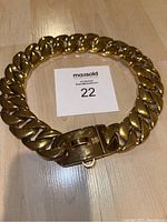 Gold-coloured metal Cuban link dog collar laid flat on wooden floor showing the chunky chain links and locking clasp.