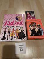 Top view photo showing all three items: RuPaul's Drag Race Paper Doll Book, RuPaul's Drag Race Card Game box, and Game of Queens card game box on a wooden floor.