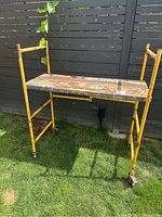 Front view of the folding baker scaffold with yellow metal frame and wooden platform, showing surface rust and weathering