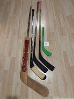 Four hockey sticks aligned on a floor, showing different sizes and conditions. The largest is a wooden Sherwood goalie stick, followed by other brands including Bauer and CCM, plus a small green foam stick.
