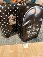 Two carry-on suitcases: one Darth Vader design by American Tourister and one black and white polka dot with cat graphic by Travelers Club, standing upright on floor.