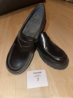 Pair of black leather Camper loafers with thick rubber soles placed on wooden floor with Maxsold number card.