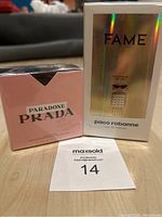 Two boxed perfumes set on a wooden floor: Fame by Paco Rabanne in a holographic box and Paradoxe by Prada in a pink box.