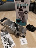 Full view of the Pro Series Mandoline Slicer with the box, instruction manual, and safety hand guard, showing the stainless steel body and handle.