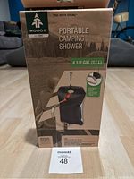 Photo of boxed Woods portable camping shower showing front of packaging with product image, brand, capacity, and accessories listed.