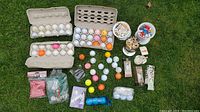 Wide view showing multiple cartons of golf balls, buckets of golf tees, and miscellaneous golf accessories laid out on grass.