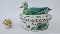 Full view of Chinese Famille Rose oval soup tureen with detailed painted foliage, lotus flowers, insects, and butterfly motifs, topped with a ceramic duck lid painted in green, blue, white, and orange.