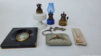 Photo showing all items in the lot from angle 1, highlighting the three mini oil lamps, the portrait print, magnifying glass with chain, silver belt buckle and enamel spoon