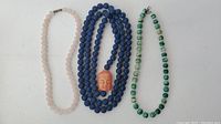 Three necklaces laid flat showing entire length and different bead types including a carved face pendant on the blue wooden necklace.