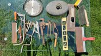 Photo of assorted hand tools including saw blades, wrenches, pliers, screwdrivers, carpenter's level, hand sanders, and a metal file, arranged on glass surface outdoors.